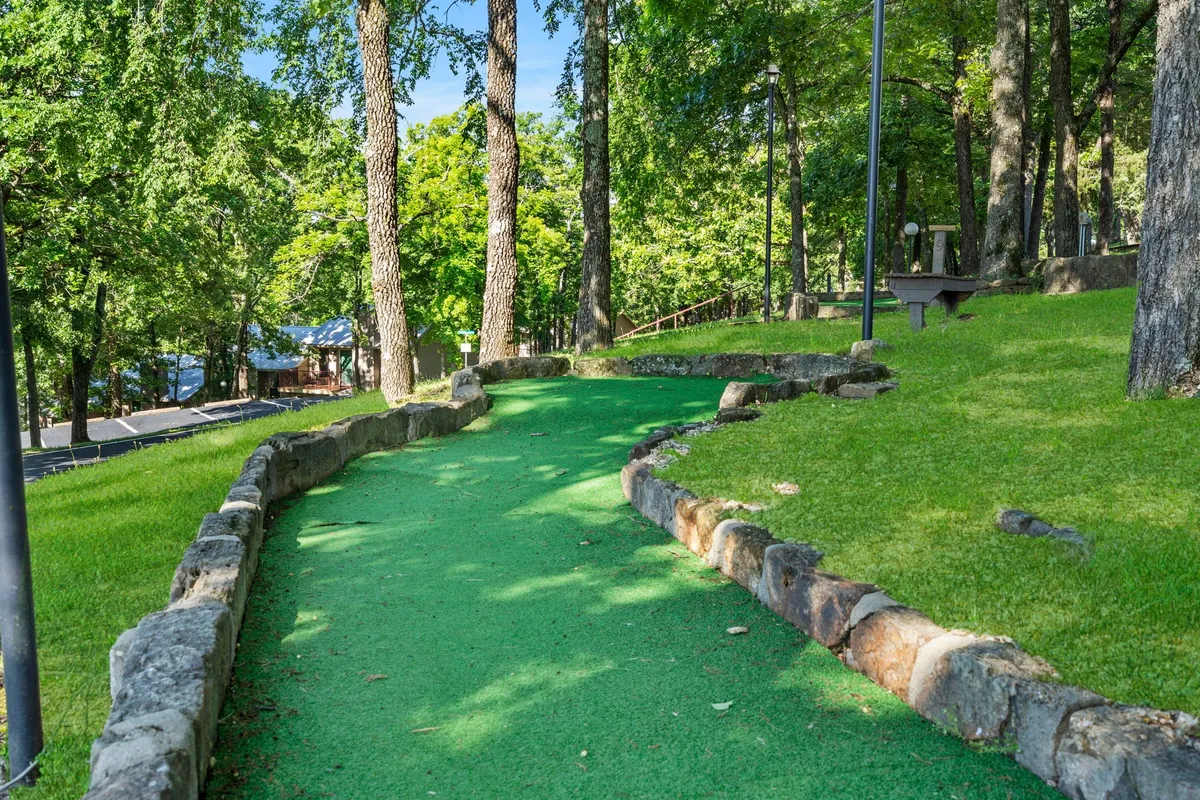 A playful mini-golf course winds through green turf, bordered by stone edges and shaded by tall trees. The area feels lively yet natural, inviting families and groups to enjoy friendly competition out in the fresh air.