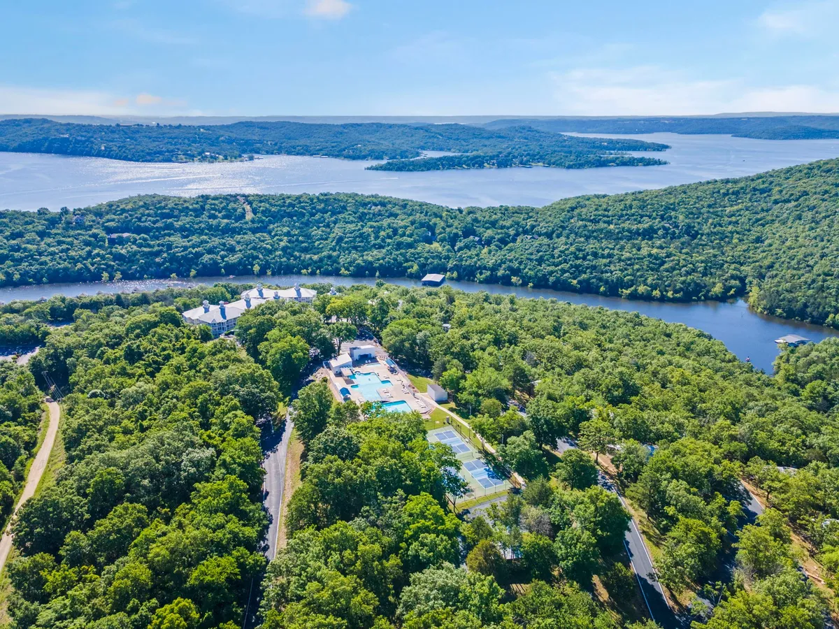 A sweeping view captures the lake framed by lush forest, with buildings and amenities like a swimming pool and tennis courts in the foreground. The pool gleams in the sunlight and the courts are ready for play, while sunset colors reflect off the water. The scene invites guests to imagine themselves swimming, playing sports, or strolling down to the lakeside.