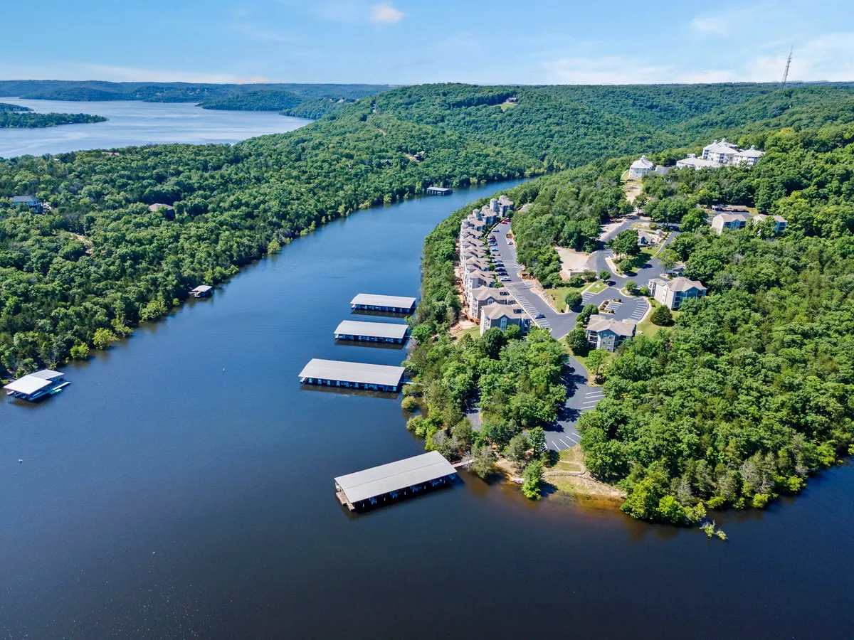 This exterior image captures a serene river with a marina, surrounded by lush forest and residential buildings. The water is central, with docked boats likely present, conveying a peaceful lakeside atmosphere ideal for boating and fishing. The overall composition highlights easy water access, tranquil views, and year-round recreation.