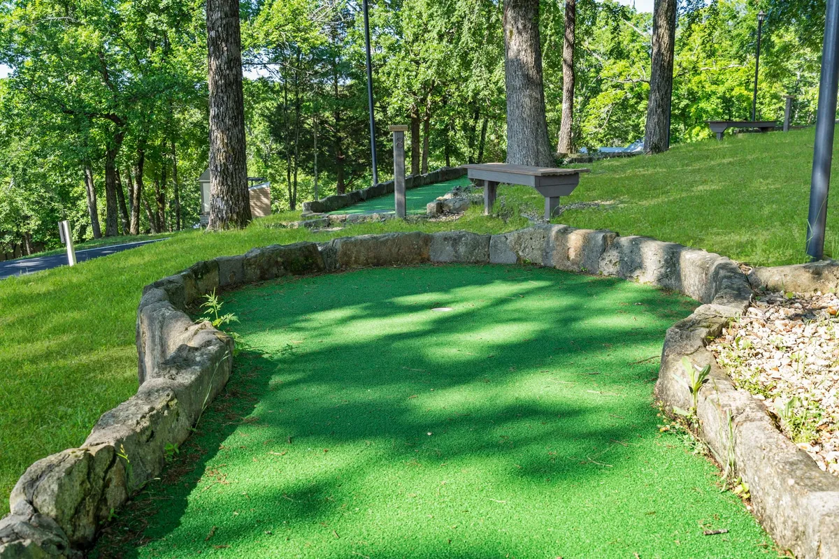 A playful mini golf course nestled among tall trees, bordered by stone, and surrounded by greenery. The scene radiates an energetic, outdoor vibe perfect for family fun and friendly competition under the sun.