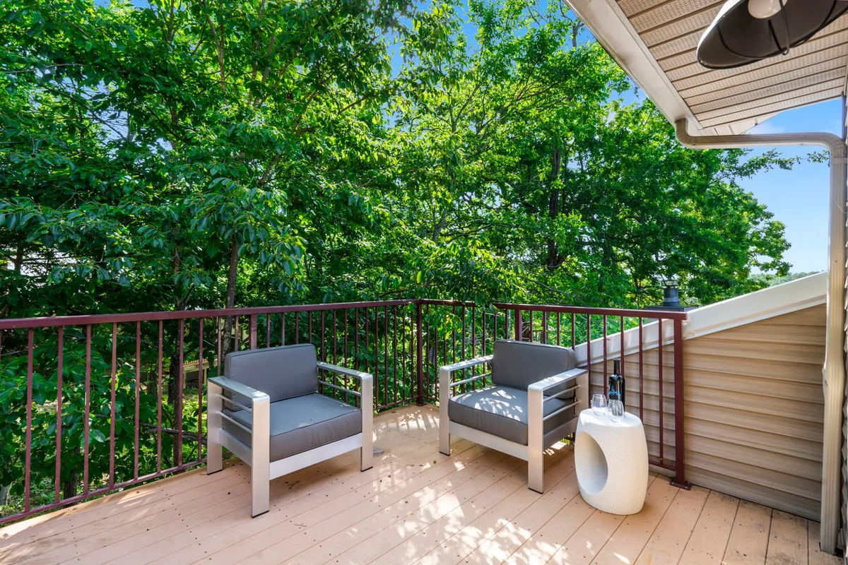 The image showcases an inviting exterior patio space featuring two modern chairs and a round accent table, surrounded by lush greenery. The peaceful setting is perfect for relaxing, enjoying a quiet morning coffee, or unwinding with a good book while soaking in the sounds and sights of nature. The setup suggests an ideal spot for intimate conversation or watching beautiful sunsets over the Ozarks.