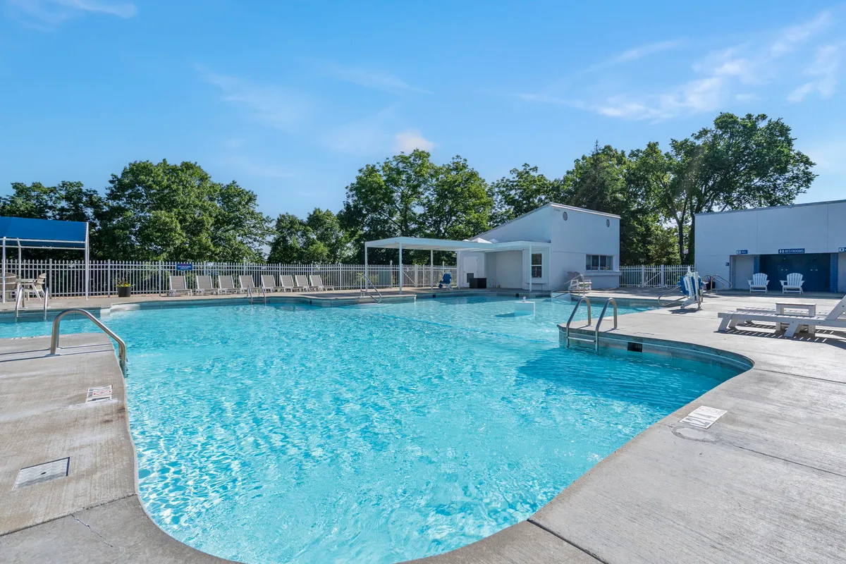 A vibrant outdoor swimming pool scene with inviting blue water, surrounded by lounge chairs and a poolside cabana. The pool is set against a backdrop of lush greenery, suggesting a relaxing atmosphere perfect for warm-weather activities like swimming, sunbathing, and socializing.